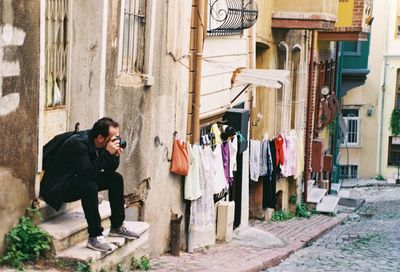 Full length of man photographing while sitting at entrance of house in city