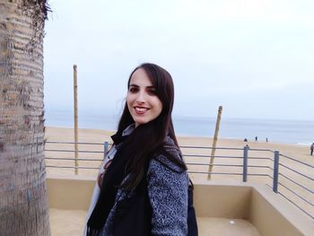 Portrait of smiling young woman standing at promenade by sea against sky