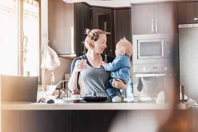 Happy mother and little infant baby boy together making pancakes for breakfast in domestic kitchen
