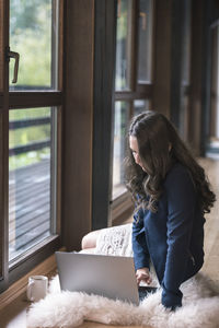 Woman using laptop while sitting on fur by window at home