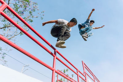 From below fearless male friends jumping above metal railing in city while performing parkour stunt on sunny day