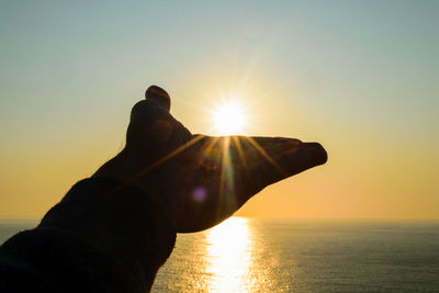 Cropped hand of man reaching sun over sea against sky