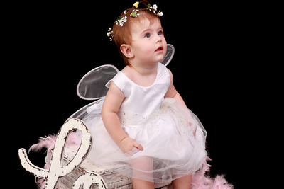 Cute baby girl wearing fairy costume sitting on box against black background