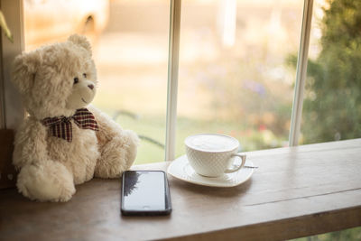 High angle view of coffee cup with mobile phone and teddy bear on table by window