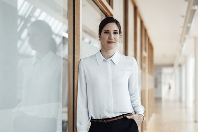 Businesswoman with hand in pocket leaning by wall in office corridor