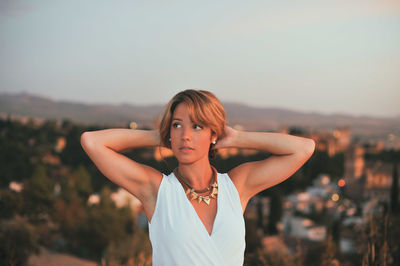 Beautiful young woman with hands behind head against city