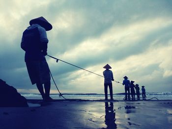 Rear view of men fishing at beach against cloudy sky