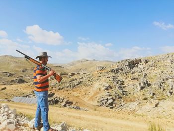 Man holding hat standing on mountain against sky