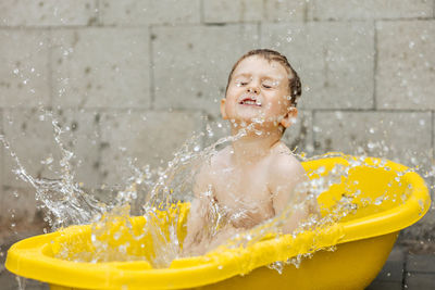Cute little boy bathing in yellow tub outdoors. happy child is splashing, playing with water  