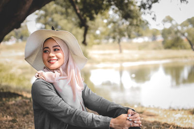Portrait of young woman wearing hat standing against lake