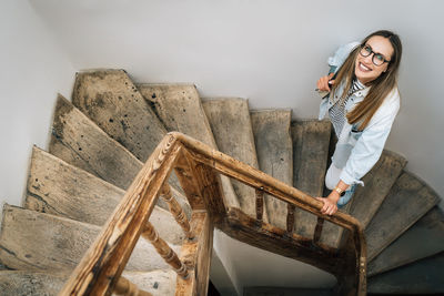 A woman with glasses smiling looks up climbing an old wooden staircase