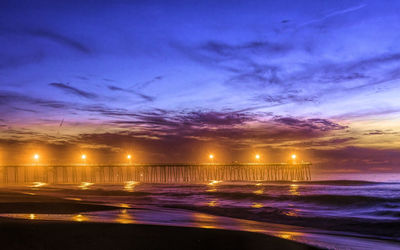 Illuminated street by sea against sky at night