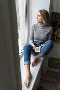 Woman wear headphones, using cellphone, sitting on windowsill listening to podcast or club house