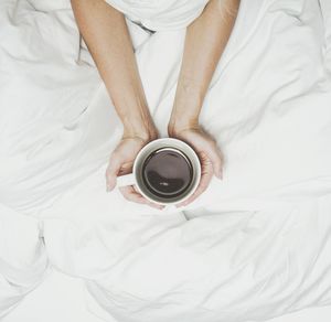 Low section of woman holding coffee cup on bed