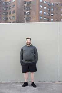 Full length portrait of overweight man with hands in pockets standing against wall in city