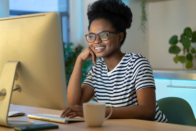 Joyful happy african american woman sitting at table watching movie and drinking tea at home