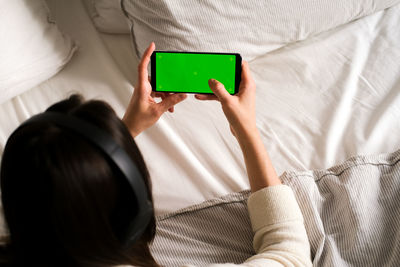 A woman with headphones lies and holds a black mobile phone with a green screen upright