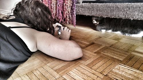 Woman photographing cat under sofa while lying on hardwood floor at home