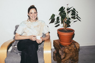 Portrait of smiling woman holding tea cup while sitting on chair by plant at home