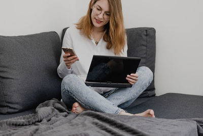 Attractive young woman working from home - female entrepreneur sitting on sofa with laptop computer and checking cell phone from comfort of home