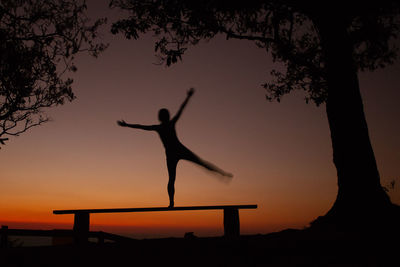 Silhouette woman balancing on bench by trees against sky during sunset