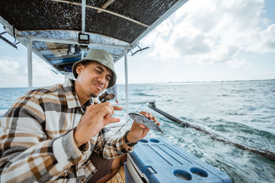 Portrait of young man drinking water while sitting on sea against sky