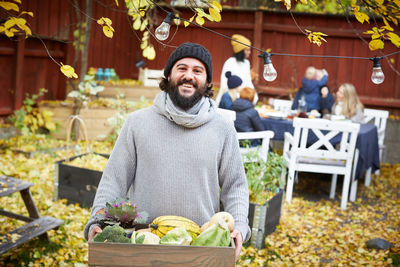 Portrait of happy man standing with vegetable basket while family and friends sitting at table in yard