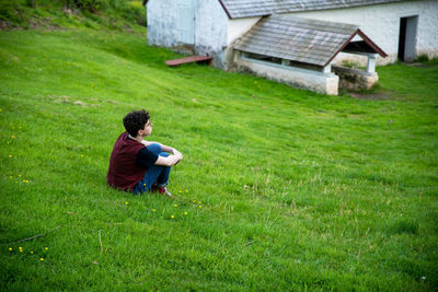 Rear view of solitary young man sitting on field against historic farm building
