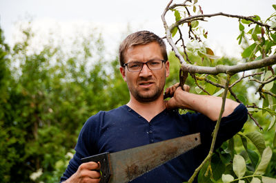 Portrait of man holding hand saw standing against plants
