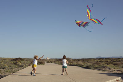 Full length of delighted little children in shorts walking on road and smiling while launching colorful kites against cloudless blue sky on sunny summer day