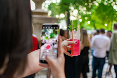 Selective focus image of woman taking photos of red cocktail at a party in city.
