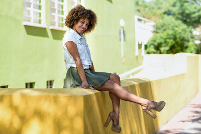 Full length portrait of smiling young woman sitting on retaining wall