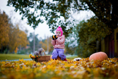 Cute girl sitting on pumpkin over autumn leaves against trees at public park
