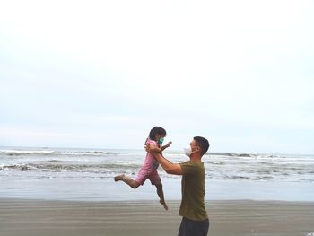 Side view of father wearing mask holding daughter at beach against sky