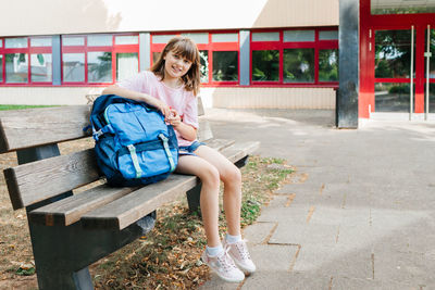 Back to school. a schoolgirl teenager sits on a bench next to a schoolbag in front of the school