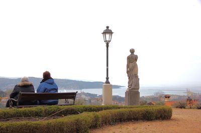 Rear view of man and woman sitting on bench over looking sea against clear sky