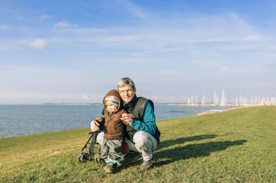 Grandfather holding granddaughter standing on grassy field against sea