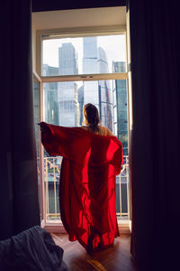 Brunette woman in a red bathrobe stands at a large window in an apartment opposite a skyscraper