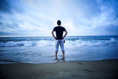 Rear view of man with hands on hip standing at beach against cloudy sky during sunset