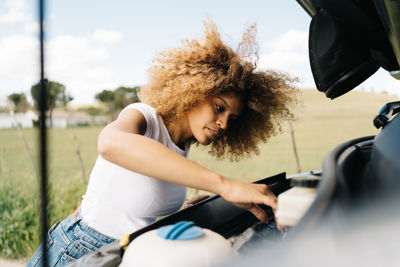 Side view of traveling female looking under opened hood of camper van while having problems during trip through summer nature