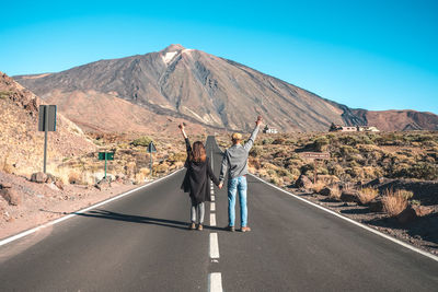 Rear view of people standing on road by mountain against sky