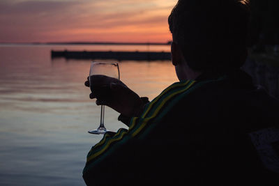 Man drinking wine by sea against sky during sunset