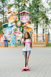 A beautiful smiling girl rides a scooter along the path of an amusement park on a summer day