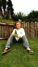 Boy holding bottle while sitting on grass against sky