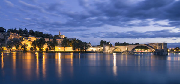 Bridge over river by illuminated buildings against sky at dusk