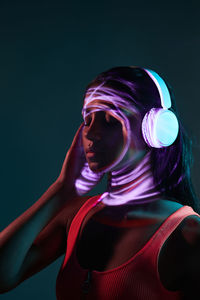 Charming brazilian female listening to music in wireless headphones while standing with eyes closed in obscure studio on dark background with shadow of light on face