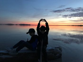 Silhouette boy making heart shape by friend sitting on driftwood at beach during sunset