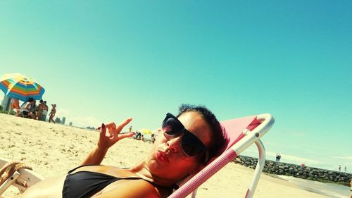 Portrait of young woman in sunglasses relaxing on deck chair at beach against blue sky