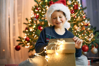 A boy in a santa hat opens a gift box next to a garlanded christmas tree. gifts for children.