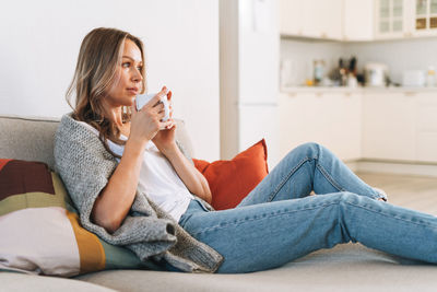 Young beautiful woman in cozy knitted grey sweater with cup of tea in hands at home